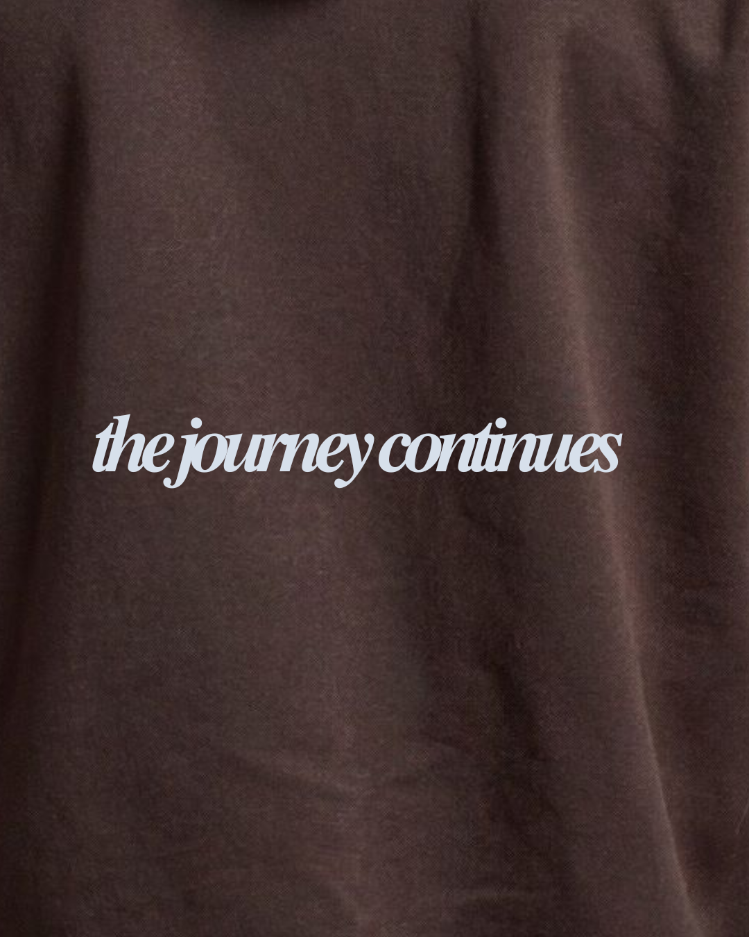 A close-up of a dark brown textured fabric, likely a t-shirt, featuring the phrase 'the journey continues' printed in a white cursive font across the center.